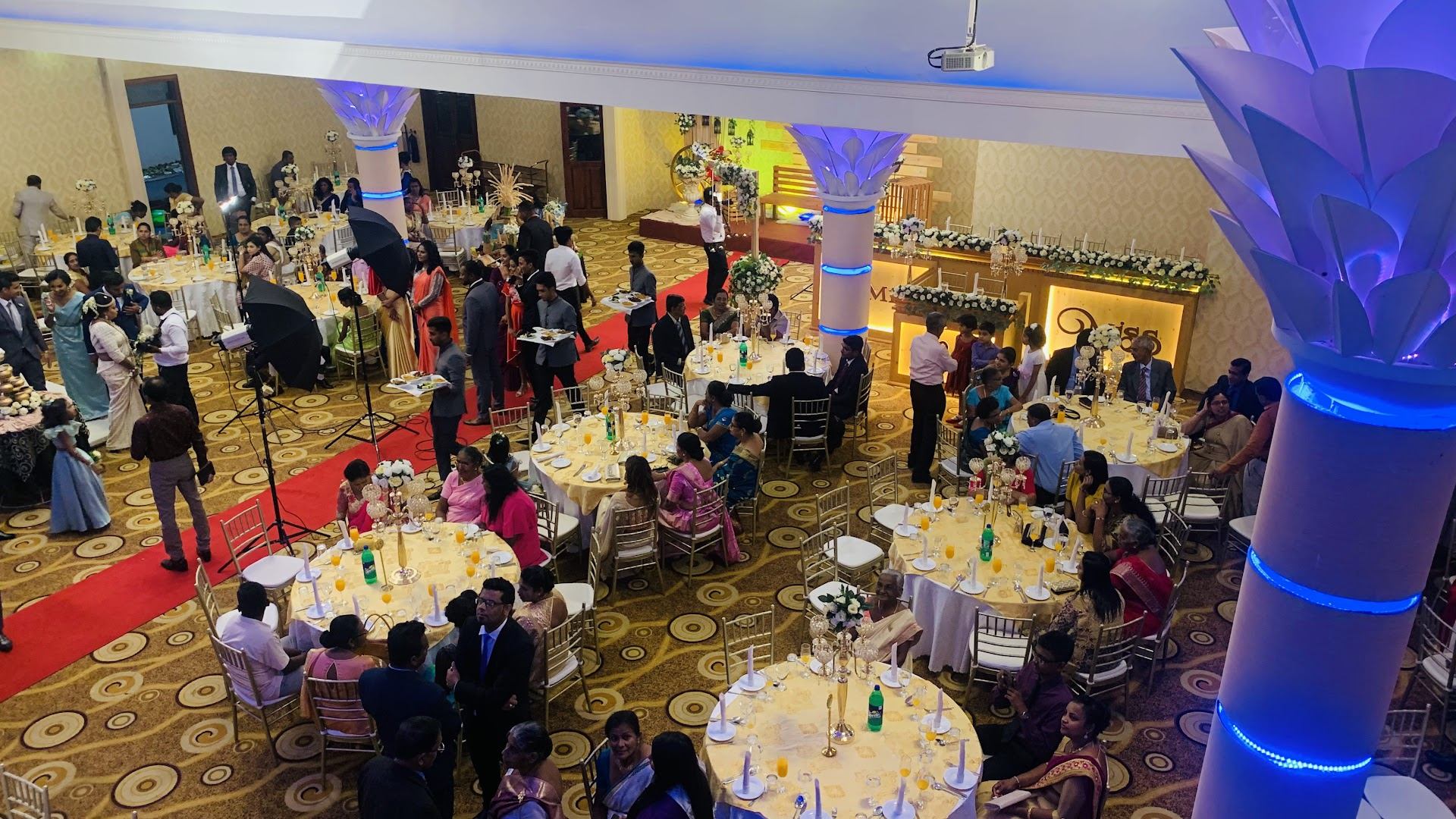 The banquet floor seen from the balcony during a wedding: round tables with gilt chairs, a red-carpet aisle and glowing lotus pillars