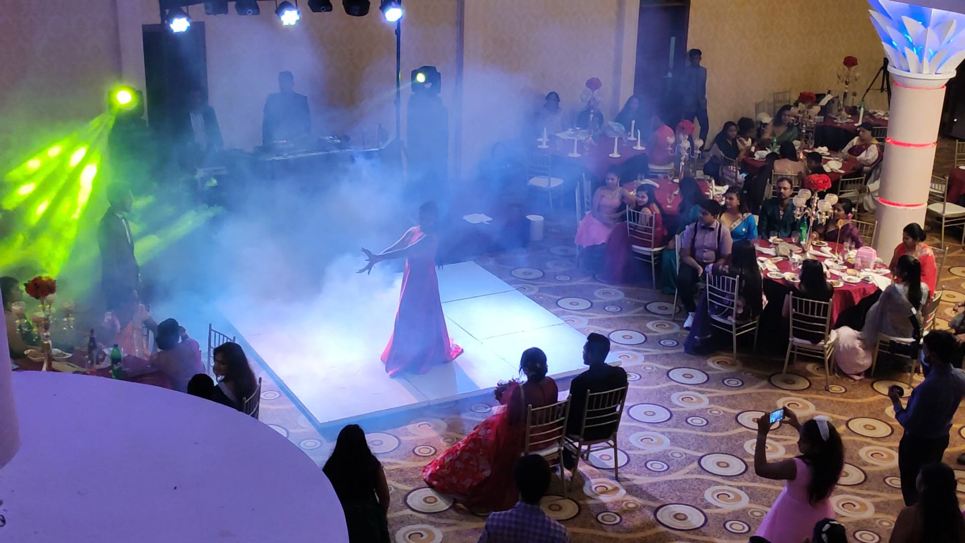 A dancer in a red gown performing in haze on a white floor, with guests watching from their tables