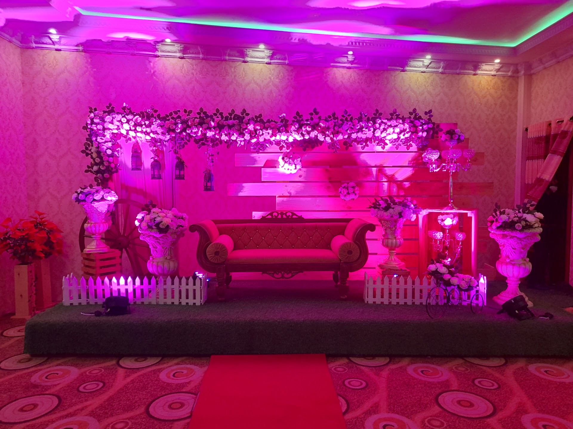A settee stage dressed with white flowers, hanging lanterns and pink light, with the red carpet leading up to it